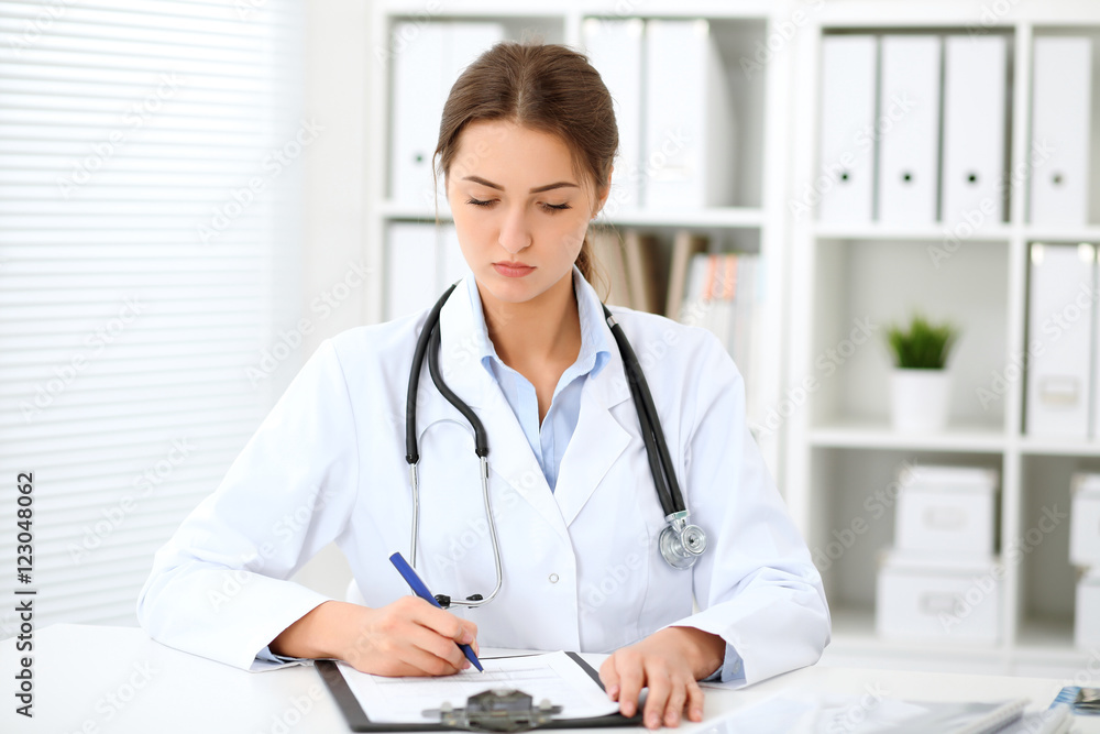 Young brunette female doctor sitting at the table and working at hospital office. Health care, insurance and help concept. Physician ready to examine patient Young brunette female doctor sitting at the table and working at hospital office. Health care, insurance and help concept. Physician ready to examine patient