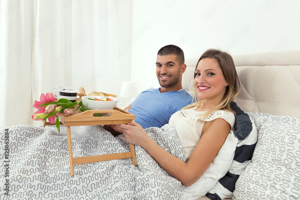 Young couple is enjoying romantic meal in a hotel room. They are lying in bed and smiling while holding a tray with a breakfast and bouquet of flowers.