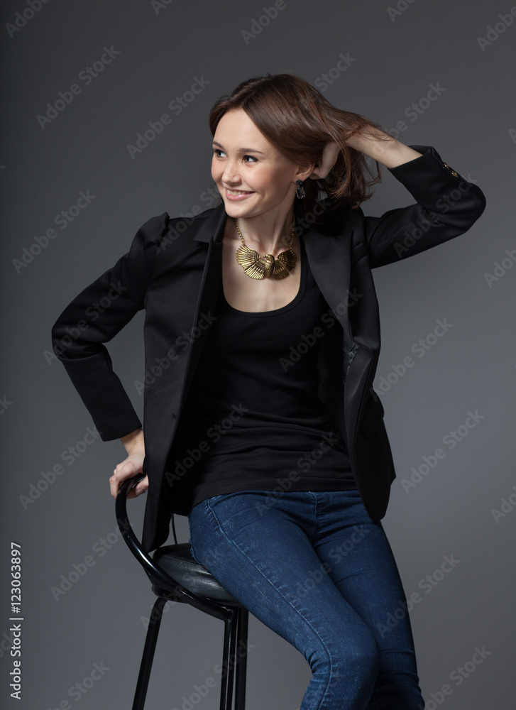 stylish young smiling woman in black blazer and dark blue jeans siting on  chair on gray background Stock Photo | Adobe Stock