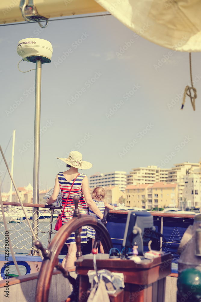 Closeup on sailing ship steering wheel and back view of mother with kids. Sunny blue sky outdoors background