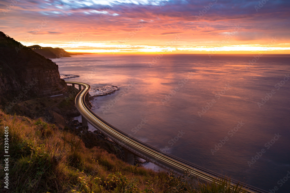 Sea Cliff Bridge on sunrise with moving traffic and dramatic beautiful sky and ocean shore on the background. The Bridge is part of NSW Grand Pacific scenic route