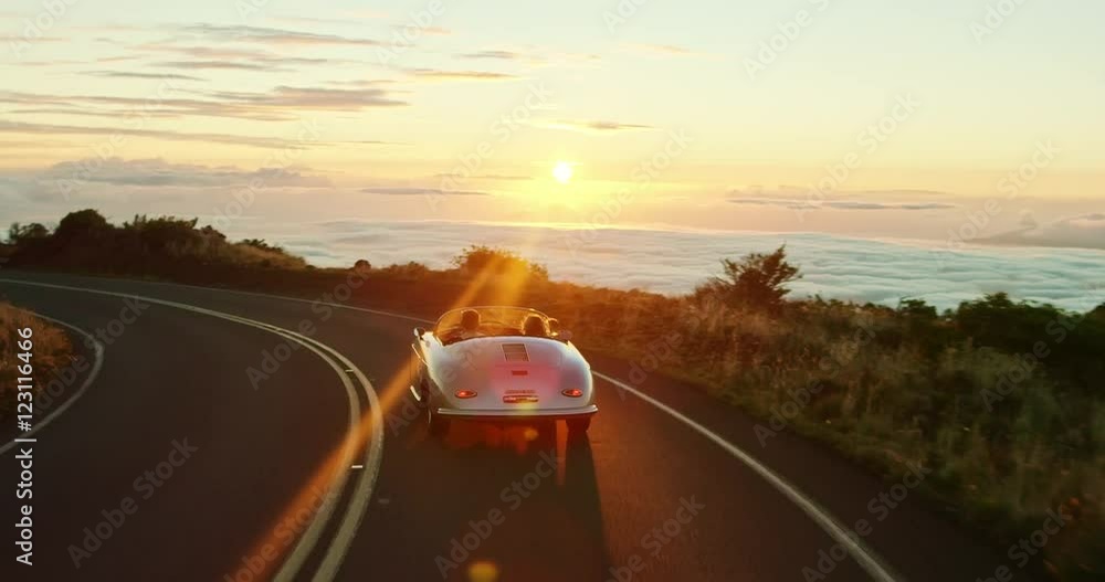 Happy couple driving on country road into the sunset in classic vintage sports car