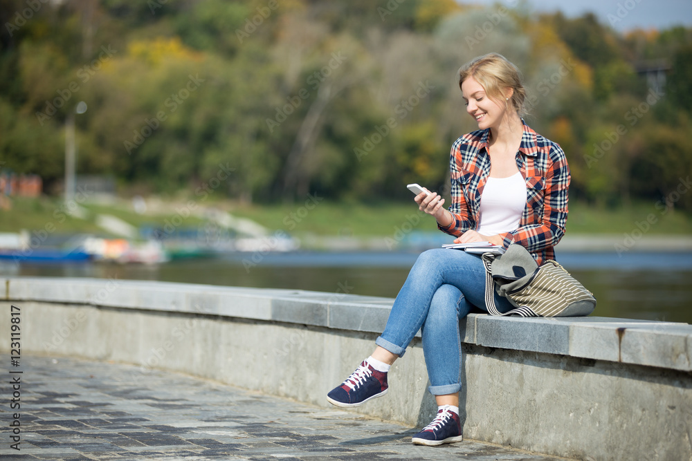 Portrait of a young attractive girl sitting her legs crossed at the bridge, waterline behind, looking at the screen of her mobile, lifestyle