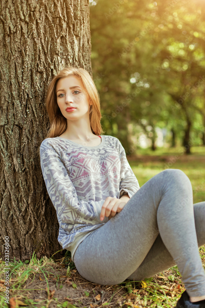 Young beautiful girl with blue eyes and blonde hair sitting near a tree Young beautiful girl with blue eyes and blonde hair sitting near a tree