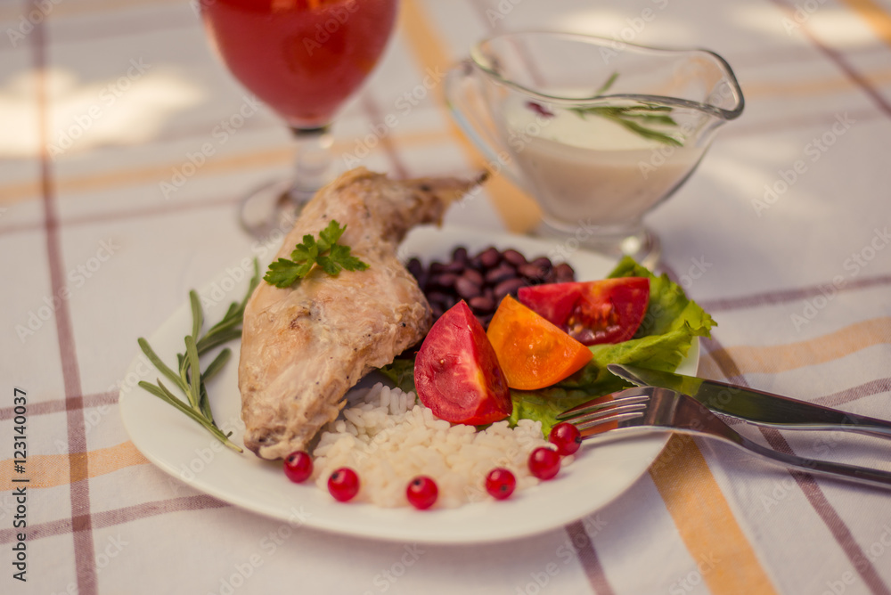 Gourmet roasted rabbit leg with rice and beans. White sauce and glass of tomato juice. Meal is served on white simply plate and checked table cloth, with cutlery. Health and light meal good for diet.