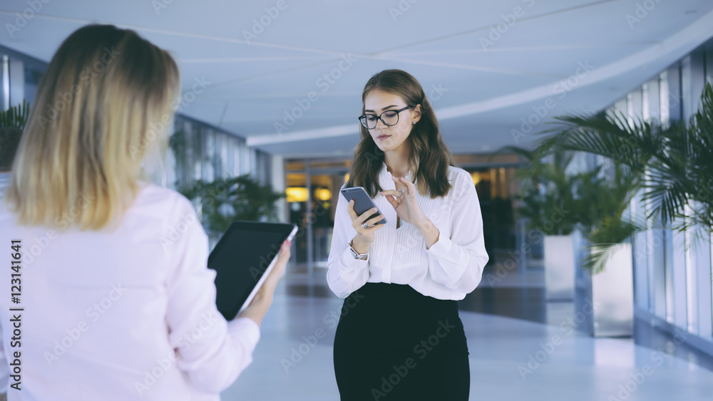Fototapeta premium Two business women standing in the lobby and checking email on the gadgets.Brunette in glasses and a white blouse holding a smartphone,in the foreground a girl with the tablet computer.