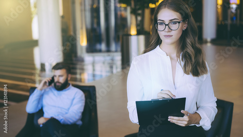 In the foreground a young business woman in a white blouse takes notes. In the hands she holding a clipboard. In the background a businessman sitting in a chair and talking on a smartphone.