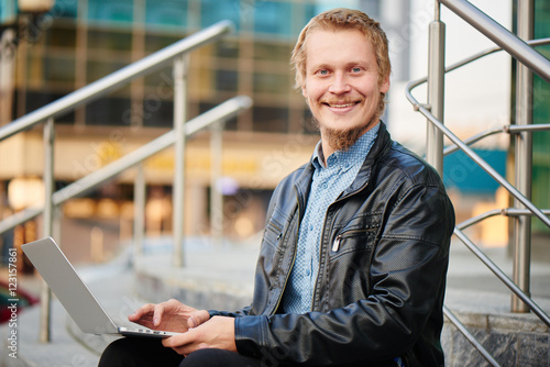 Portrait of a blond guy with a beard in a leather jacket and shirt, smiling sitting on the steps with the computer on my lap, in the background the building with a glass facade