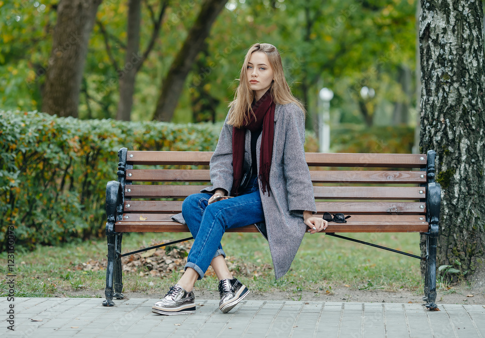 Beautiful stylish young woman in a warm scarf blue jeans trendy shoes and cozy coat sitting alone on bench waiting in fall city park Beautiful stylish young woman in a warm scarf blue jeans trendy shoes and cozy coat sitting alone on bench waiting in fall city park