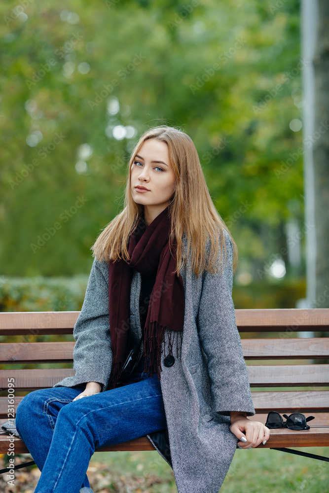 Beautiful stylish young woman in a warm scarf blue jeans trendy shoes and cozy coat sitting alone on bench waiting in fall city park Beautiful stylish young woman in a warm scarf blue jeans trendy shoes and cozy coat sitting alone on bench waiting in fall city park