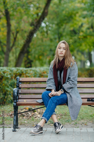 Beautiful stylish young woman in a warm scarf blue jeans trendy shoes and cozy coat sitting alone on bench waiting in fall city park