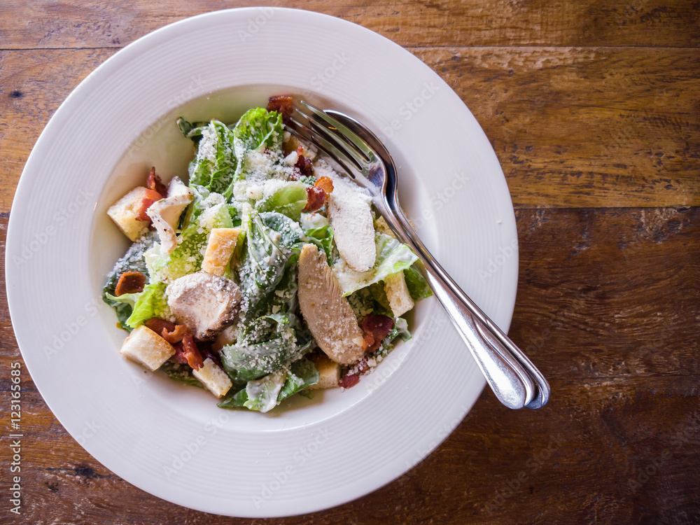 Top view of the delicious Caesar salad that contains green cos vegetable, crispy bacon, crouton and grilled chicken sprinkle over with Parmesan cheese in a white dish that is placed on a wooden table. Top view of the delicious Caesar salad that contains green cos vegetable, crispy bacon, crouton and grilled chicken sprinkle over with Parmesan cheese in a white dish that is placed on a wooden table.