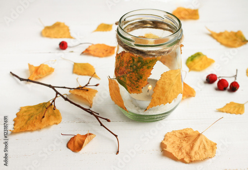 Autumn-themed DIY candle. Tealight candle in glass jar, decorated dried golden leaves. Fall time interior decor. 