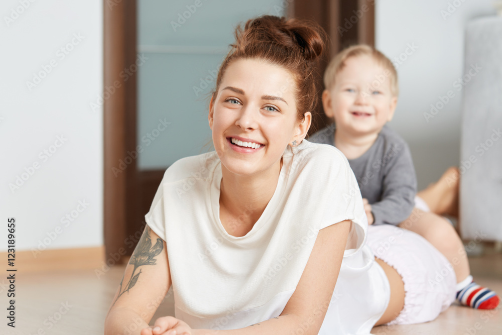 Nice family shot of young mother and her little son playing on the floor at home. Attractive Caucasian woman in white top, lying on her belly. Smiling kid straddling happily on her shinny mommy. Nice family shot of young mother and her little son playing on the floor at home. Attractive Caucasian woman in white top, lying on her belly. Smiling kid straddling happily on her shinny mommy.