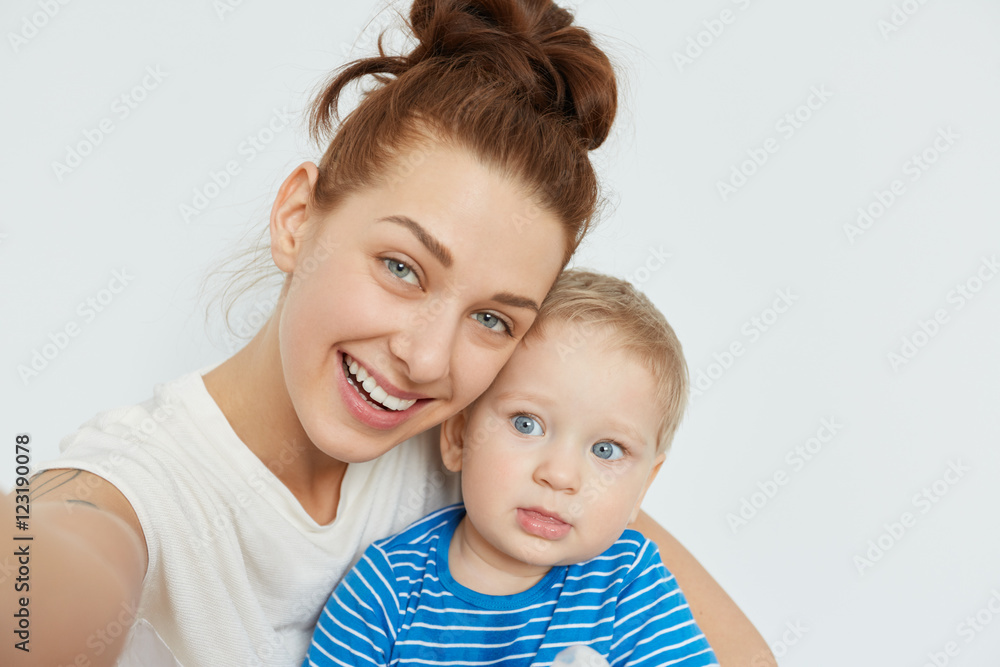 Nice selfie of mother and child on white background. Young Caucasian woman with blue eyes, smiling, holding in her arms little blond boy in blue stripped T-shirt. Happy family self-portrait. Nice selfie of mother and child on white background. Young Caucasian woman with blue eyes, smiling, holding in her arms little blond boy in blue stripped T-shirt. Happy family self-portrait.