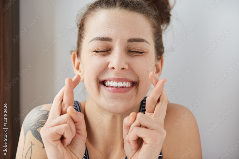 Young female Caucasian student keeping her fingers crossed and eyes closed to make a desired wish. Charming white smile, positive heartwarming look of girl sincerely believing in her victory. Young female Caucasian student keeping her fingers crossed and eyes closed to make a desired wish. Charming white smile, positive heartwarming look of girl sincerely believing in her victory.
