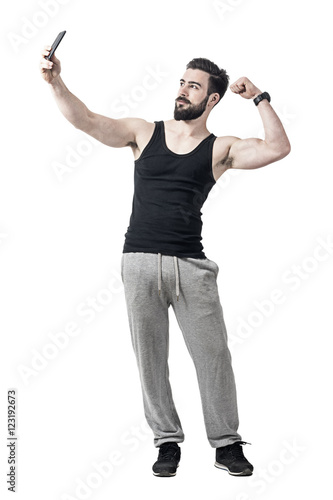 Fit young man flexing bicep arm muscle while taking selfie photo with mobile phone. Full body length portrait isolated over white studio background