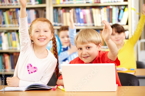 Smiling little students sitting in classroom and raising their hands to answer