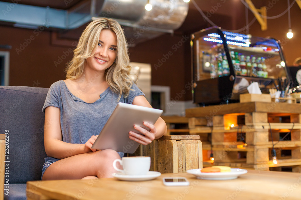 Young blonde girl with beautiful smile posing while sitting with computer in modern coffee shop interior, gorgeous cheerful female working with tablet during rest time in cozy cafe Young blonde girl with beautiful smile posing while sitting with computer in modern coffee shop interior, gorgeous cheerful female working with tablet during rest time in cozy cafe