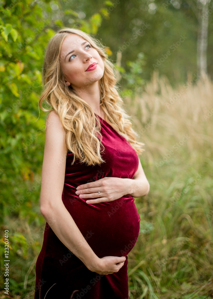 Dreamy pregnant woman in a burgundy dress with long blond hair enjoying leisure in park