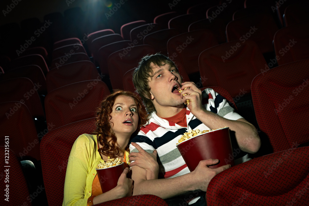 Naklejka premium Young girl sitting terrified at cinema hall, her impassive boyfriend eating popcorn
