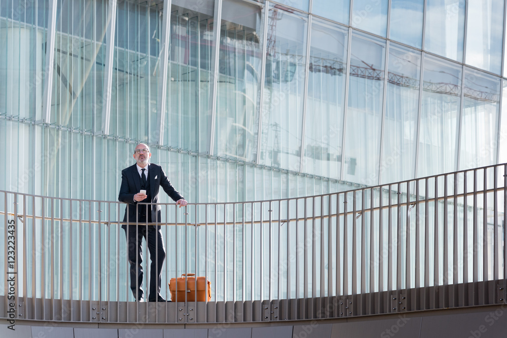 Middle-age contemporary businessman leaning on a handrail holding smart phone - work, business, technology concept Middle-age contemporary businessman leaning on a handrail holding smart phone - work, business, technology concept