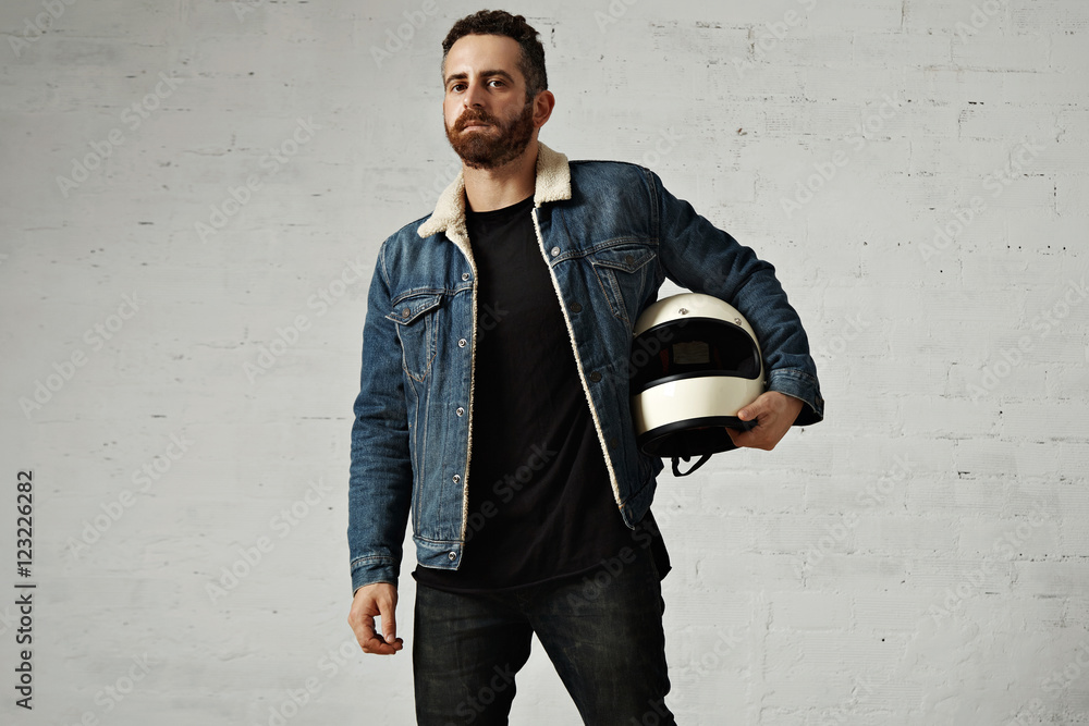 Brutal attractive motor biker wears shearling denim jacket and black blank  henley shirt, holds vintage beige motorcycle helmet, isolated in center of  white brick wall Stock Photo | Adobe Stock