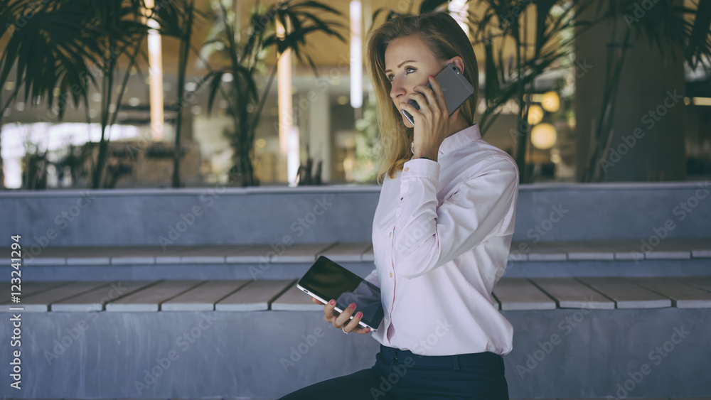 Fototapeta premium View from the side, a woman with blonde hair in a light pink shirt sitting and talking on a cell phone, in her hand she holds a tablet computer with a blank screen.Film effect, blurred background.