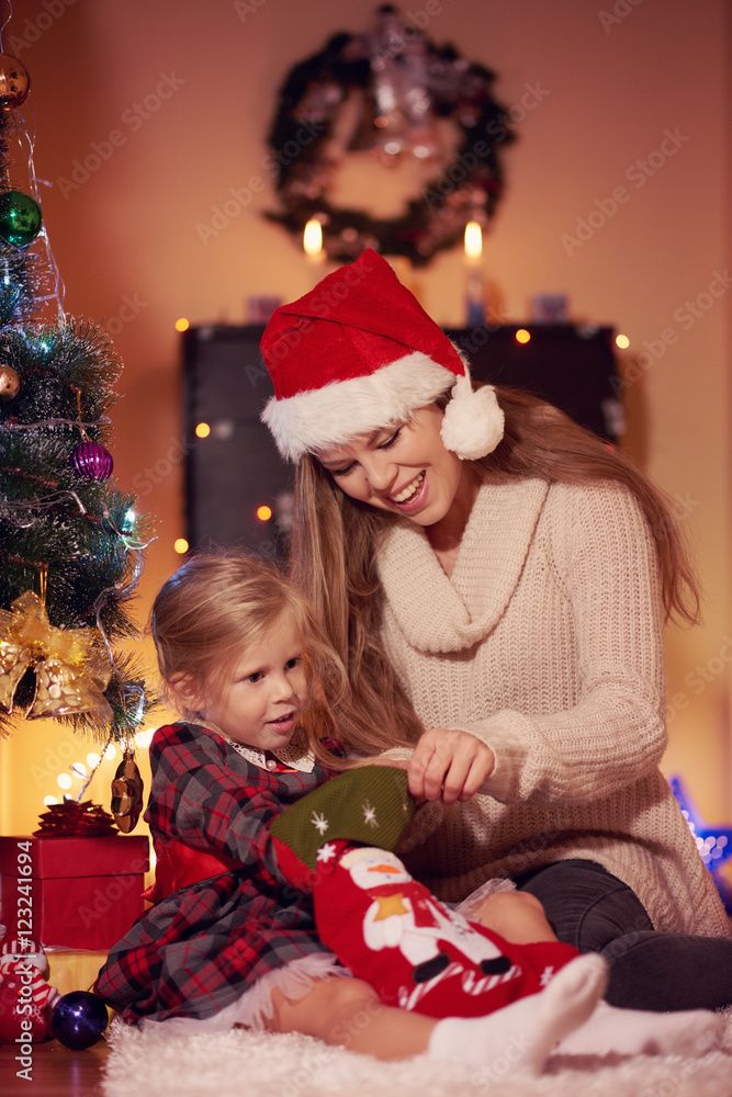 Cheerful mother in Santa hat with her little daughter wearing festive dress looking for xmas present in red sock. Happy family sitting on carpet with gifts celebrating New Year at home.