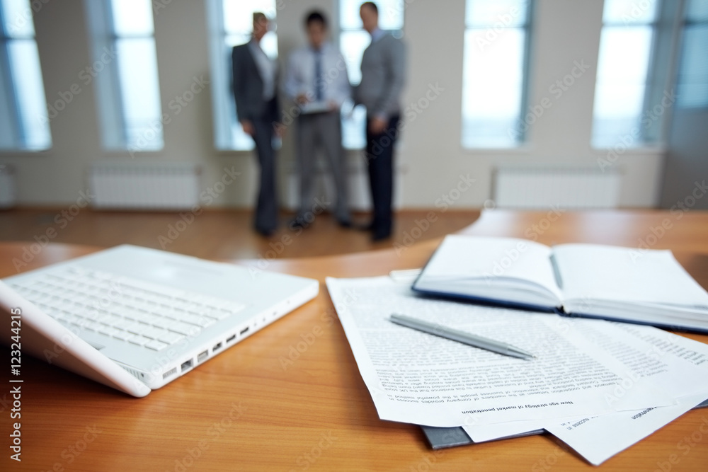 Office table with laptop and documents, three business people are in the background Office table with laptop and documents, three business people are in the background