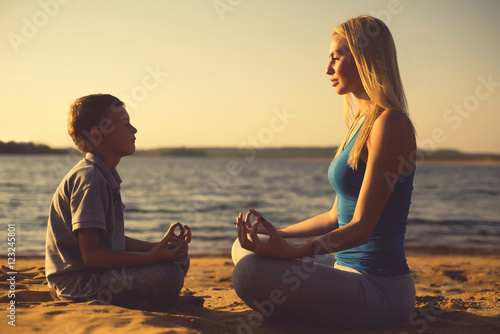 Toned photo. Mother and son do yoga by the water on the sand. Mom teaches son to meditate.