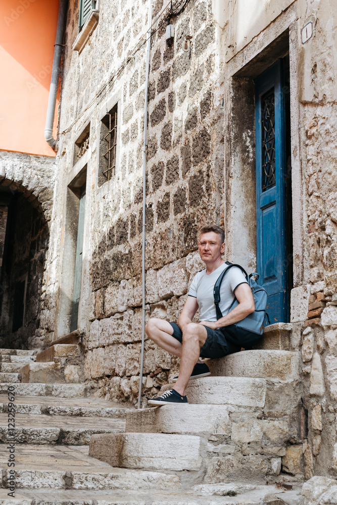 Stylish tourist. Man dressed in a white shirt and blue shorts with a blue backpack over his shoulder. Sitting on the steps of an old European city