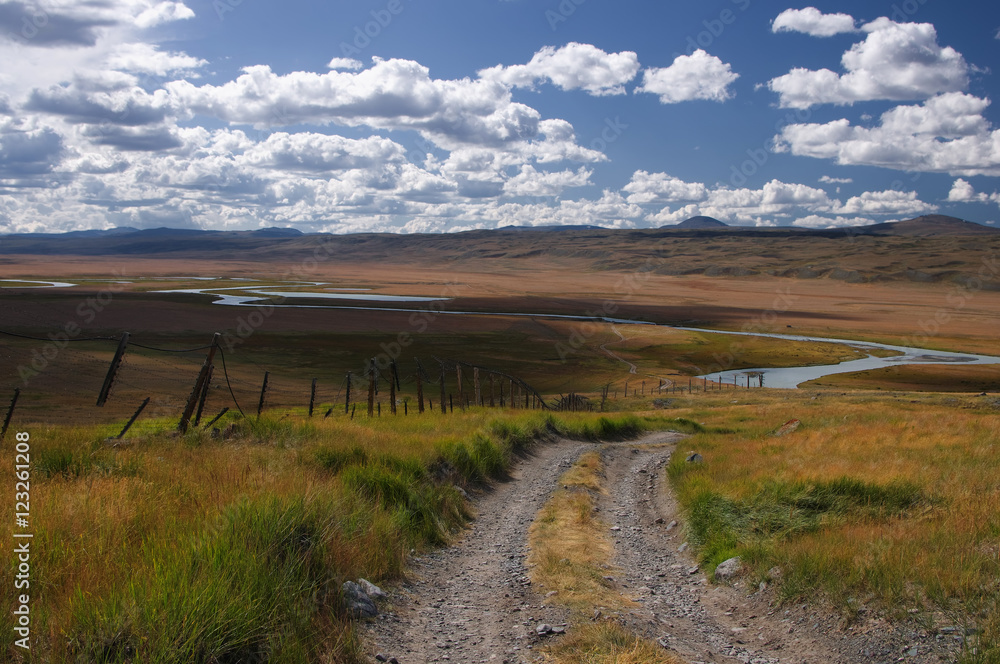 Road along the border line on a highland mountain plateau with the green grass at the background of the valley of white river under a blue sky with white clouds, Plateau Ukok, Altai, Siberia, Russia