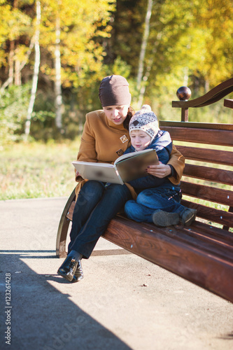 Young woman and little boy sitting on the bench in autumn Park and reading book. Family enjoying time together. Mother teaches son