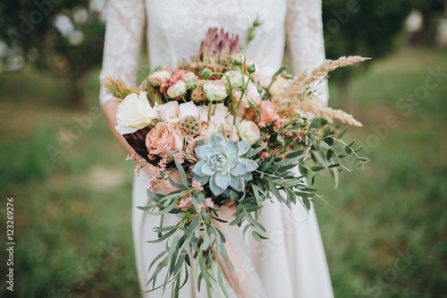 bride in a dress standing in a green garden and holding a wedding bouquet of flowers and greenery