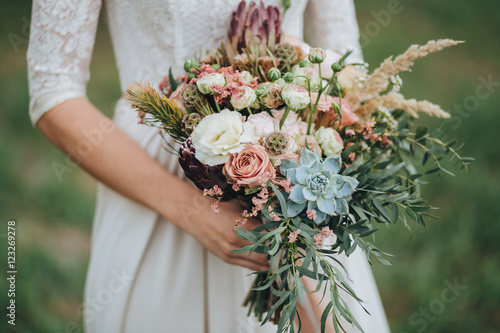 bride in a dress standing in a green garden and holding a wedding bouquet of flowers and greenery