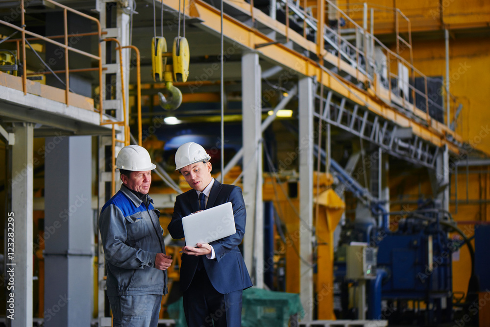 The man in the suit and the helmet holds the portable computer and shows up on the screen to the worker in overalls in an industrial building The man in the suit and the helmet holds the portable computer and shows up on the screen to the worker in overalls in an industrial building