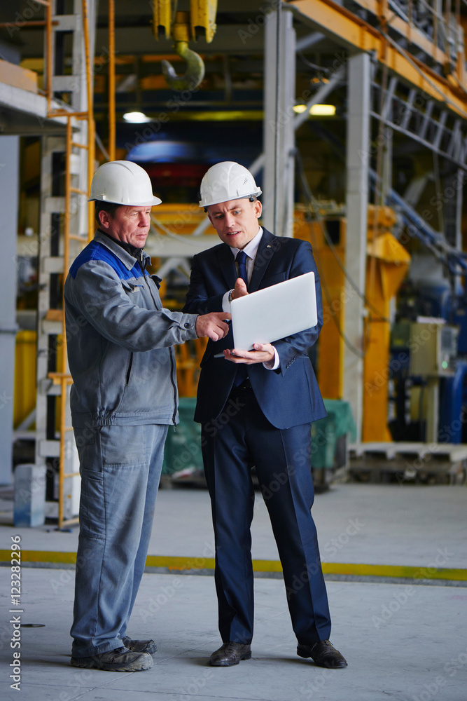 The man in the suit and the helmet holds the portable computer and shows up on the screen to the worker in overalls in an industrial building The man in the suit and the helmet holds the portable computer and shows up on the screen to the worker in overalls in an industrial building