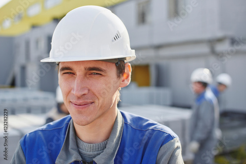 Portrait of a Builder in overalls and helmet on the background of a group of people around a cart with a brick © amixstudio