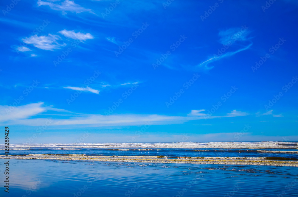 Pacific northwest ocean beach shoreline.  Coastal salt water splashing on shore.  Wispy white clouds and vibrant blue sky reflecting on wet sands.