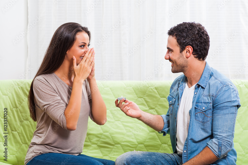 Young couple is sitting at sofa at home. Man is proposing to his woman.