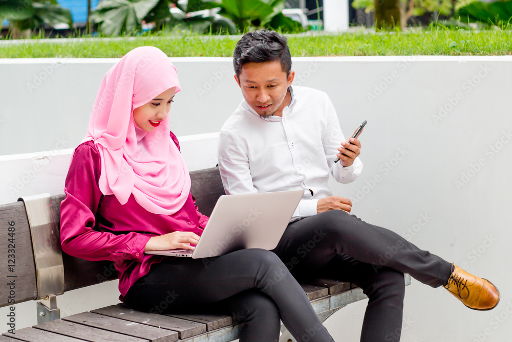 Beautiful young Asian Muslim business woman wearing head scarf (hijab) with a laptop outside the office discussing with young guy with a smartphone. Beautiful young Asian Muslim business woman wearing head scarf (hijab) with a laptop outside the office discussing with young guy with a smartphone.