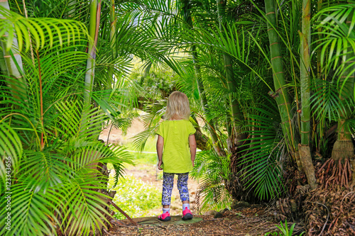 Little blond girl staying among palm trees outdoors