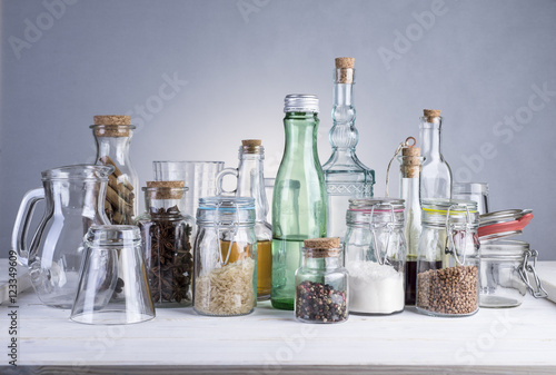 Still life of transparent glass bottles, cans and glasses on a white wooden table.