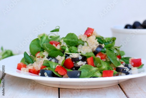 Fresh healthy vegan salad with quinoa, corn salad, black olives, red pepper and olive oil on white plate cloth on white wooden background