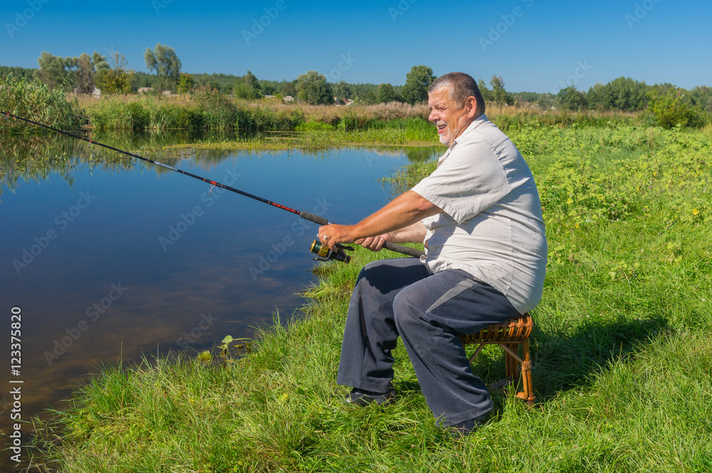 Senior fisherman sitting on a wicker stool with spinning rod and ready to catch fish in small river Merla in central Ukraine Senior fisherman sitting on a wicker stool with spinning rod and ready to catch fish in small river Merla in central Ukraine