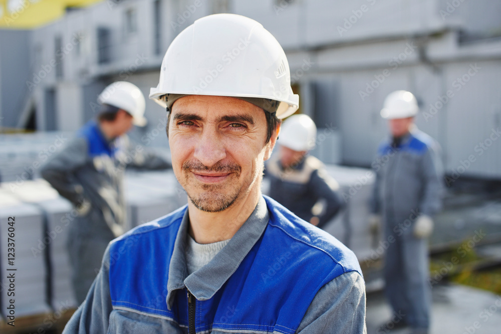 Portrait of a Builder in overalls and helmet on the background of a group of people around a cart with a brick Portrait of a Builder in overalls and helmet on the background of a group of people around a cart with a brick