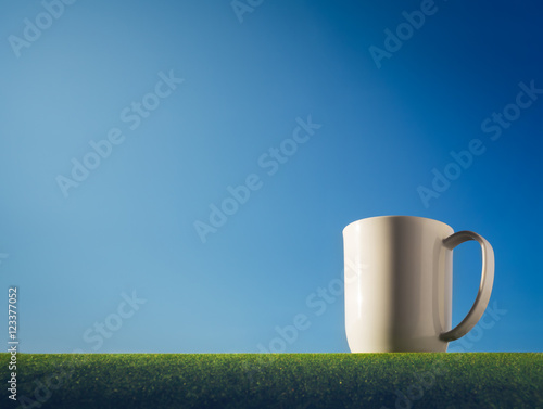 Morning mug coffee with blue sky background and grass floor.