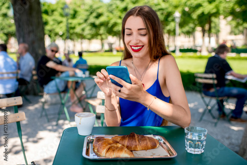 Beautiful woman in the blue dress sitting at the cafe with coffee and croissant using mobile phone outdoors at the park.