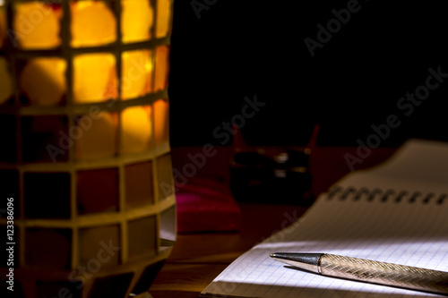 Pen over a graph notebook, beside a crystal pencil holder of different colors. Chiaroscuro